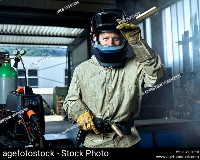 Manual worker wearing protective welding helmet holding welding machine while standing at factory