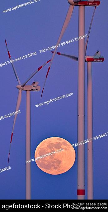 16 April 2022, Brandenburg, Petersdorf: The full moon shines at dusk between the masts of wind turbines in East Brandenburg
