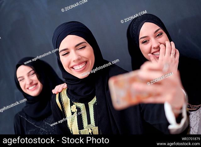 group of young beautiful muslim women in fashionable dress with hijab using mobile phone while taking selfie picture in front of black chalkboard representing...
