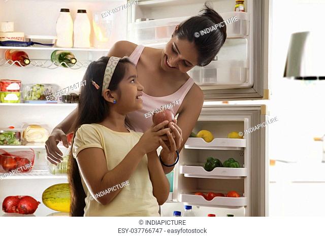 Mother giving apple to daughter in front of open refrigerator