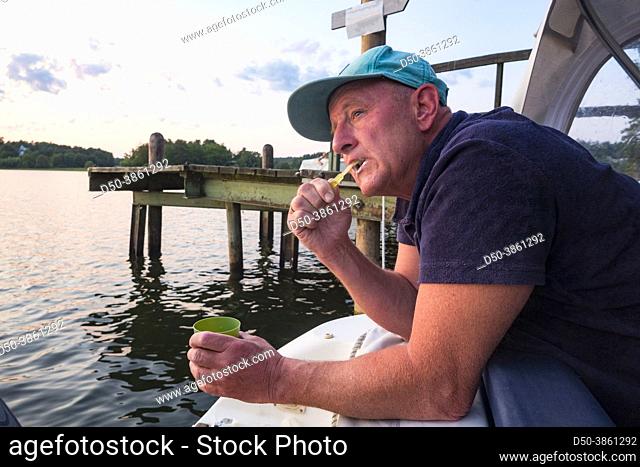 A middle-aged man brushes his teeth on the aft section of a small motorboat while camping