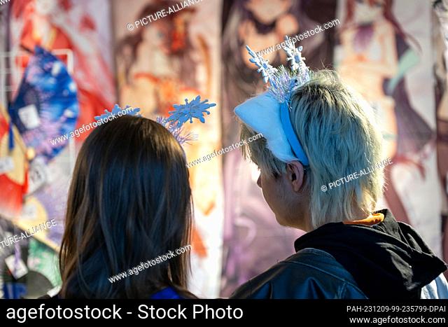 09 December 2023, Bavaria, Coburg: A woman and a man with white ears and a snowflake hairband on their heads look at manga merchandise