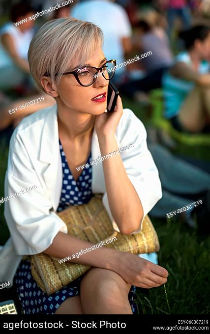Blonde girl in glasses talks on a cell phone on the blurry background outdoors. She sits on the grass in a blue dress with white pateerns and a white cloak