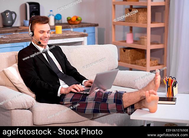 Close up view of a man working on freelance at home. Male in jacket and pyjamas trousers on couch with laptop on his knees