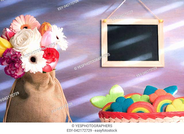 Beautiful bouquet of diverse flowers in a jute sack, a basket with multi-shaped colorful cookies and a blank blackboard hanging on the purple wall