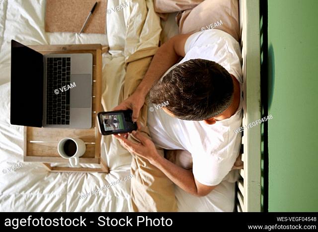 Man video chatting with friend through mobile phone while sitting by laptop in bed