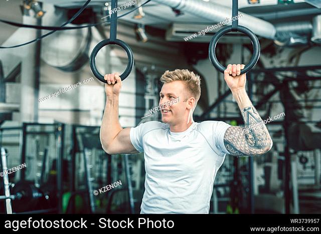Portrait of a blond handsome young man with strong muscular arms smiling while hanging on gymnastic rings at the gym