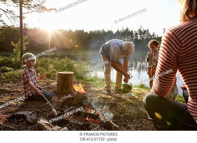 Grandparents and grandchildren at campfire at sunny lakeside in woods