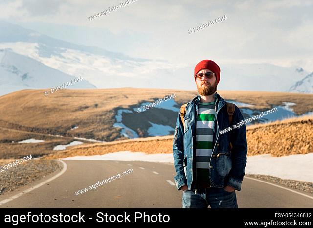 A stylish bearded hipster in sunglasses with a vintage backpack stands on a country road asphalt on a sunny day. The concept of hitchhiking and hiking