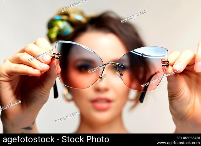 Portrait of young woman with sunglasses in hand. Female is looking at the camera through glasses. Focus on sunglasses and fingers, face is in bokeh