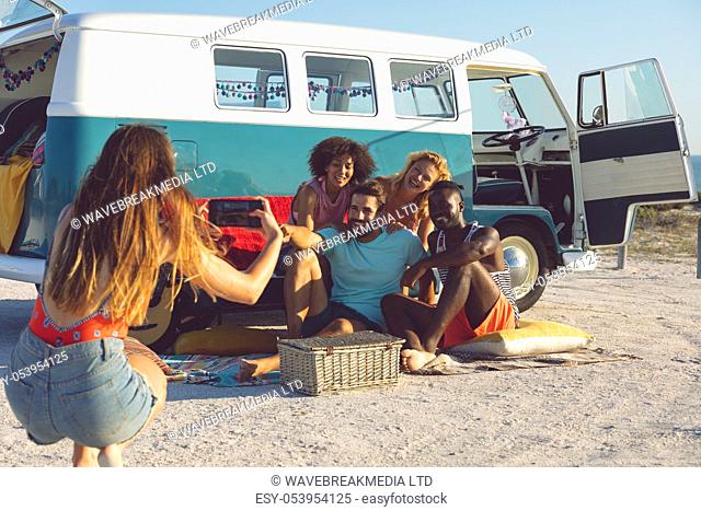 Rear view of young Caucasian woman taking pictures of her friends with mobile phone near camper van at beach