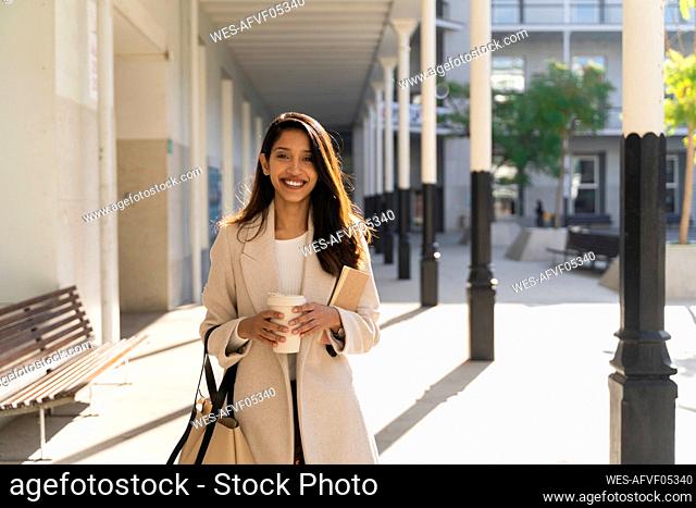 Portrait of smiling young woman with book and takeaway coffee in the city
