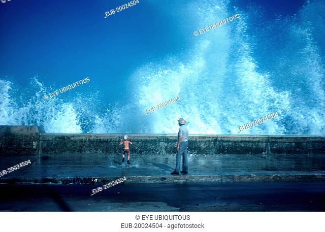 Old man and small boy standing on pavement with waves crashing over the sea wall