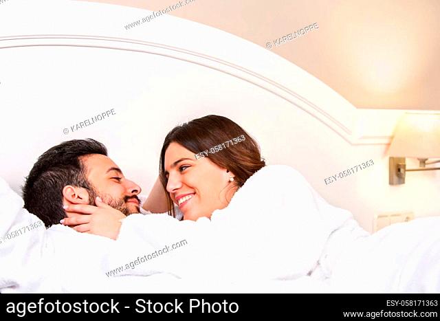 Close up portrait of cute young couple in bathrobe. Laying together on bed in hotel room. Girl showing affection