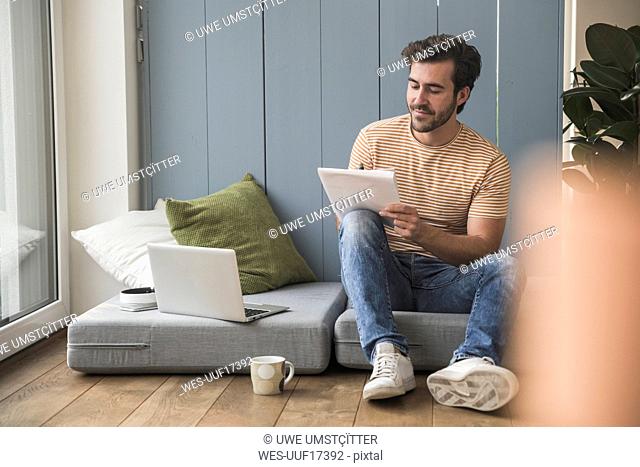 Young man sitting on mattress, using laptop, taking notes