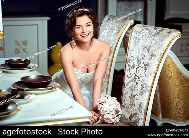 Beautiful young bride in fashionable gown with floral bouquet sitting near dinner table. Interior photo