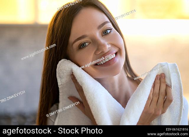 Tenderness. Close up of young pretty woman with toothy smile looking at camera touching her face with towel indoors