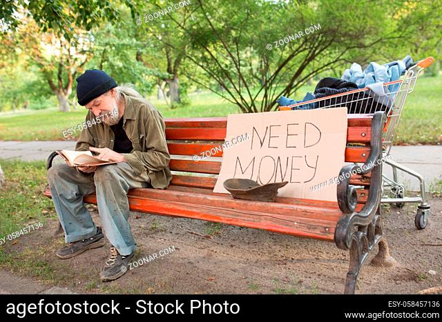 View of tramp in old dirty clothes sitting on bench reading a book. On bench old homeless beggar and board with sign need money