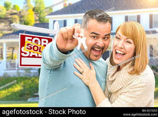 Happy mixed-race couple with keys in front of sold for sale real estate sign and new house