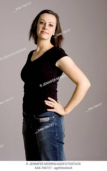 Woman in her early thirties stands confidently, hands on hips, wearing black t-shirt and blue jeans, looking at camera with slight smile