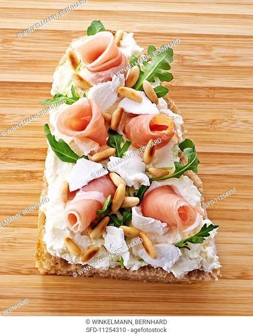 A slice of bread topped with cream cheese, prosciutto, rocket and pinenuts