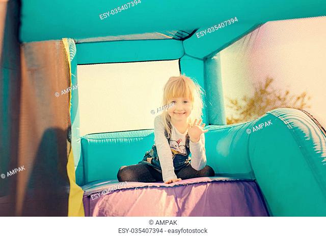 Single little happy blond girl dressed in blue jeans skirt getting ready to go down inflatable purple and green outdoor slide