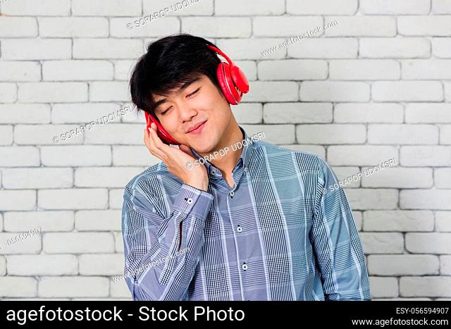 Portrait of Asian handsome young man he smile listening to music in headphones on brick wall background. Happy teenage guy smiling