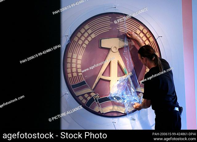09 June 2020, Saxony-Anhalt, Marienborn: Carpenter Gisbert Peuker removes the protective film from a cover of a GDR emblem that comes from the concrete stele at...