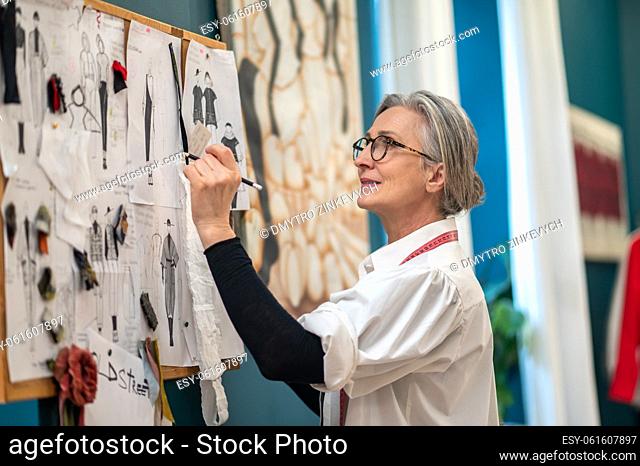 Fashion designer. Encouraged gray-haired woman in glasses with measure on neck standing sideways to camera drawing sketch on wall in atelier