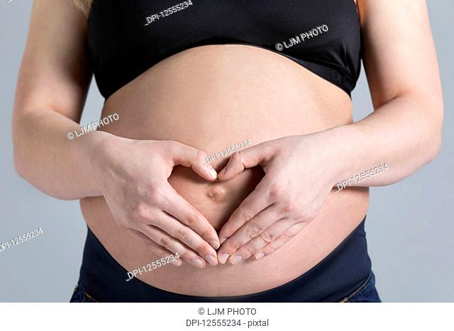 A young pregnant woman holding her belly in a studio and making a heart shape over her unborn child; Edmonton, Alberta, Canada