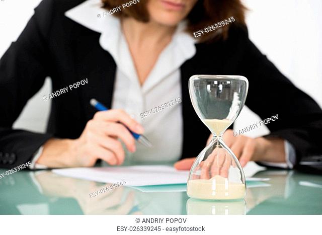 Close-up Of Businesswoman Filling Form With Hourglass On Desk