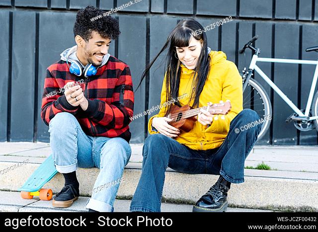 Smiling woman playing guitar while sitting by male friend on footpath