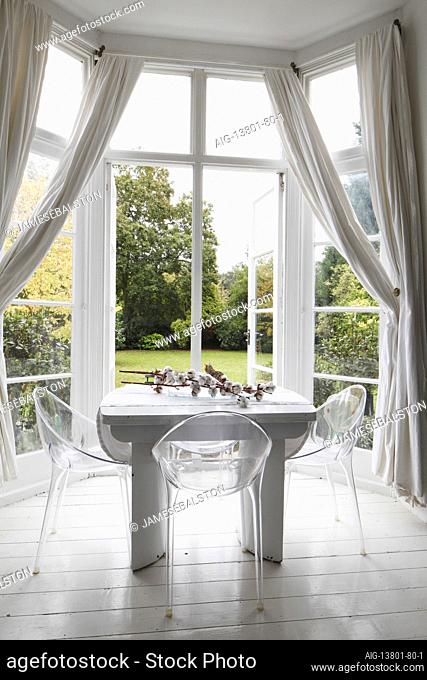 White table and perspex chairs in bay window with muslin drapes and doors open to the garden | | Designer: Amber Jeavons