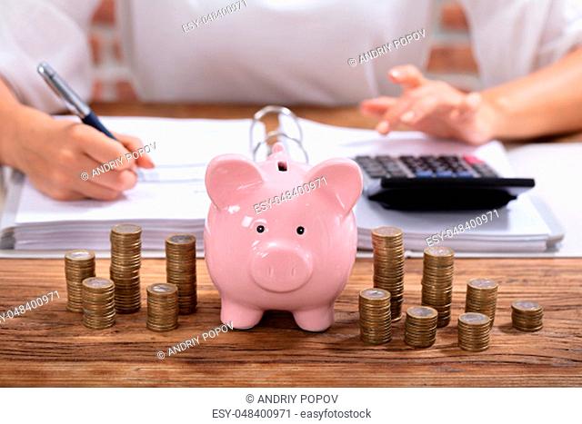 Stacked Coins And Pink Piggy Bank In Front Of Businesswoman Calculating Invoice On Wooden Desk