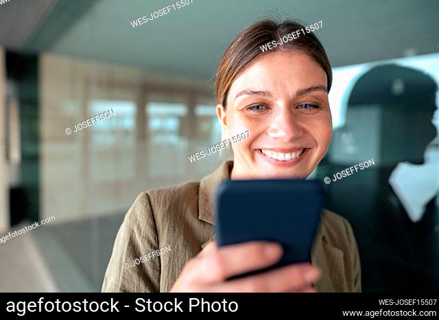 Smiling businesswoman using smart phone leaning on glass wall