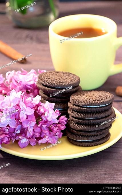 Yellow cup of tea with chocolate biscuits with a sprig of lilac and cinnamon. Tea still life yellow service, dessert and spring flowers