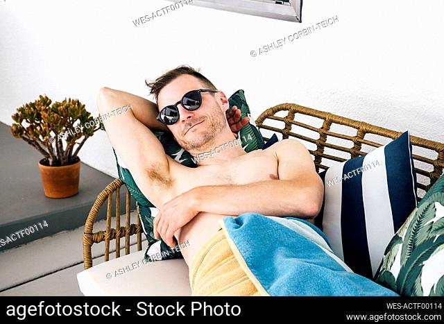 Shirtless man with hand behind head relaxing on sofa at patio
