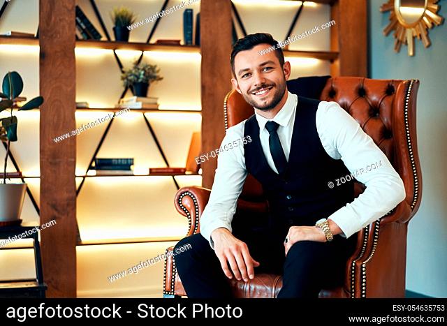 Smiling handsome man sitting and posing in armchair in modern luxury interior. Male beauty