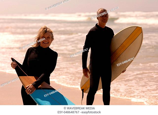 Senior couple with surfboard standing on beach and looking at camera