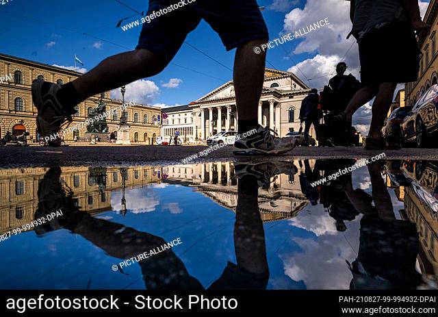 27 August 2021, Bavaria, Munich: The Bavarian State Opera is reflected in a puddle on Maximilianstrasse in the evening. On the left is the equestrian monument...