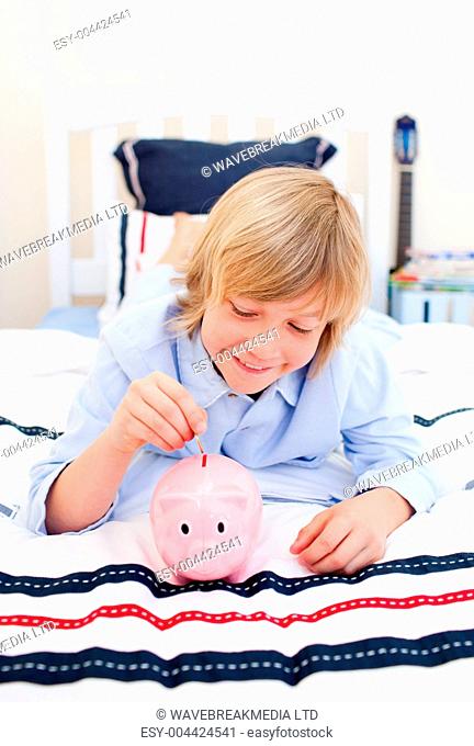 Cute boy inserting a coin in a piggybank lying down on bed