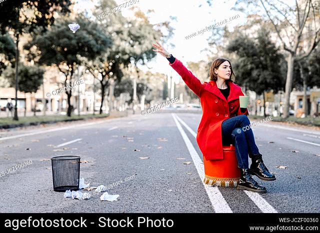 Young woman throwing crumpled paper in garbage can while sitting on street