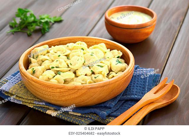 Cooked tortellini stuffed with cheese served with parsley cream sauce in wooden bowl, photographed on dark wood with natural light (Selective Focus