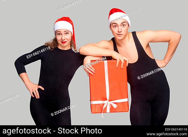 funny couple in black dress and red christmas hat holding gift box together and looking at camera. Indoor studio shot, isolated on gray background