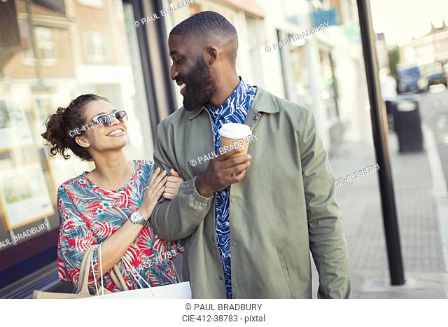 Smiling young couple with coffee walking arm in arm along storefronts
