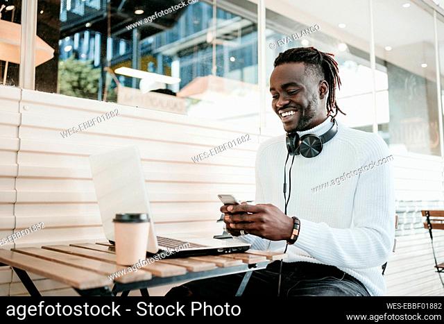 Smiling creative businessman with laptop using mobile phone in cafe