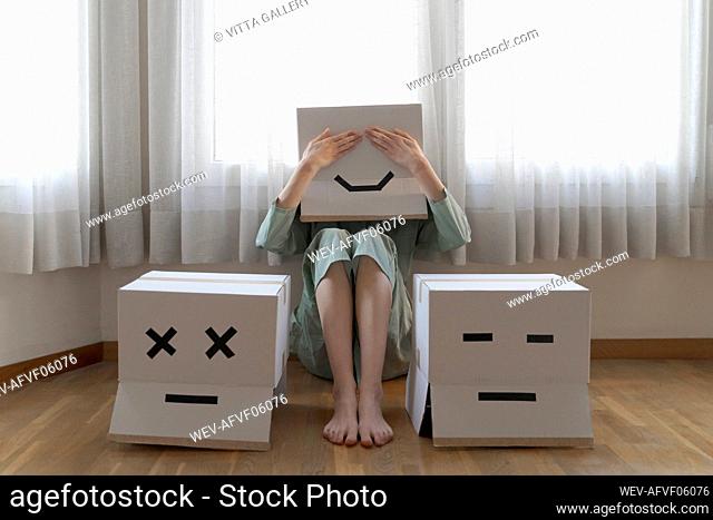 Woman wearing a cardbox on head with bored smiley sitting on floor in front of laptop, covering the eyes