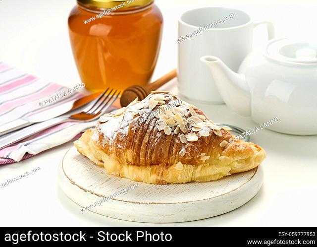 Baked crisp croissant is sprinkled with sugar powder and almond flakes on a wooden board, white ceramic brew and a cup on a white table. Breakfast
