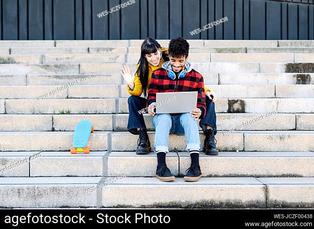 Smiling woman waving hand on video call through laptop while sitting with friend on steps