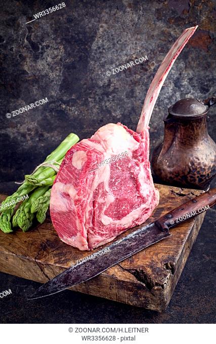 Raw dry aged Wagyu Tomahawk Steak with green asparagus as close-up on old cutting board